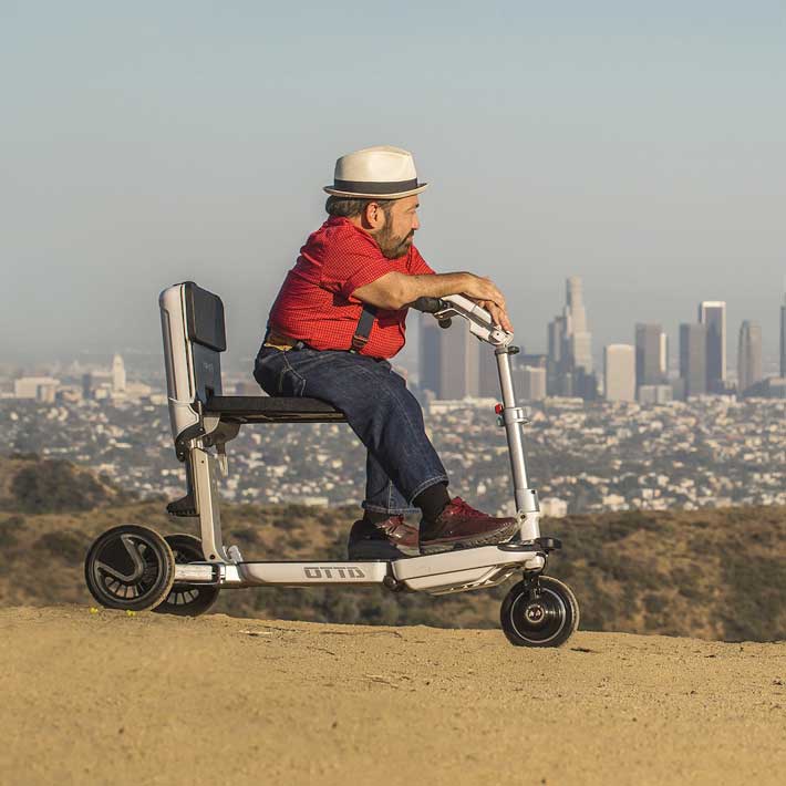 Mann fährt silbernen ATTO Scooter auf Anhöhe mit Stadtpanorama, Seitenansicht fahrbereit